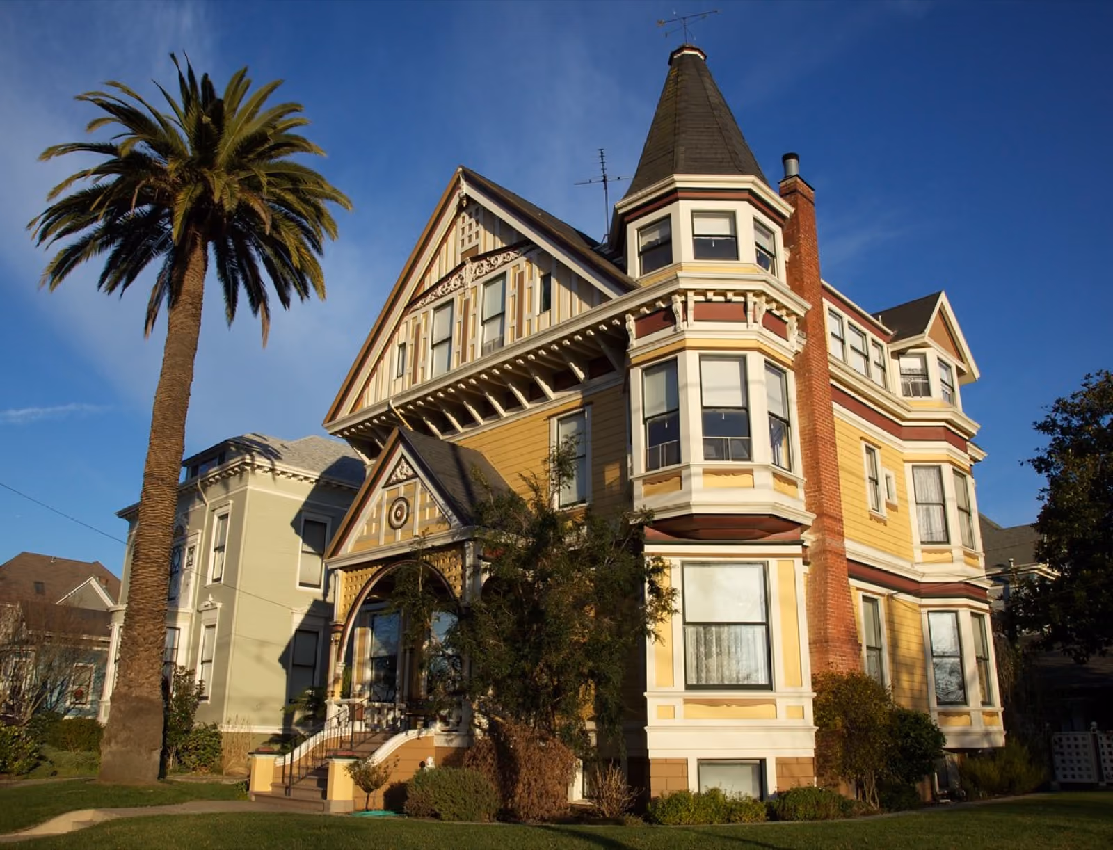 A tall Queen Anne Victorian in yellow and red, a palm rising beside its corner tower.