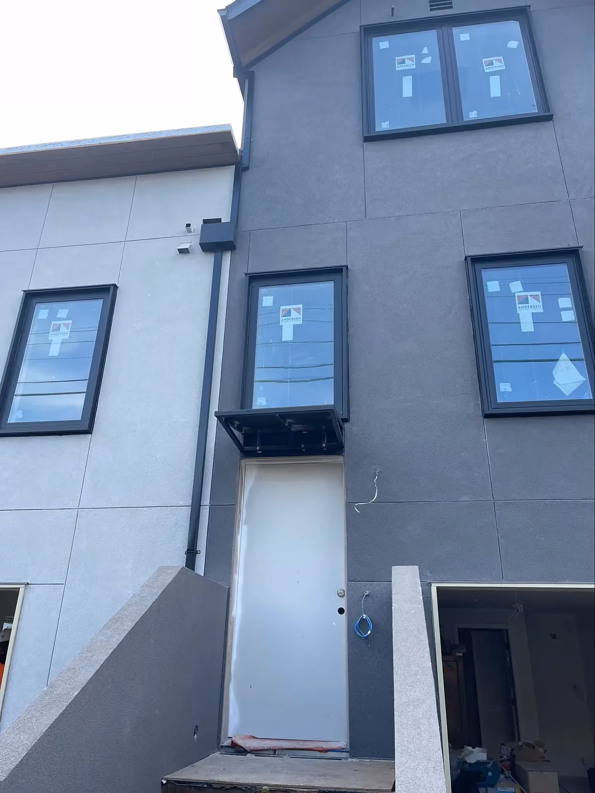 A black downspout running down the corner of a new stucco townhouse, the windows still carrying their factory stickers.
