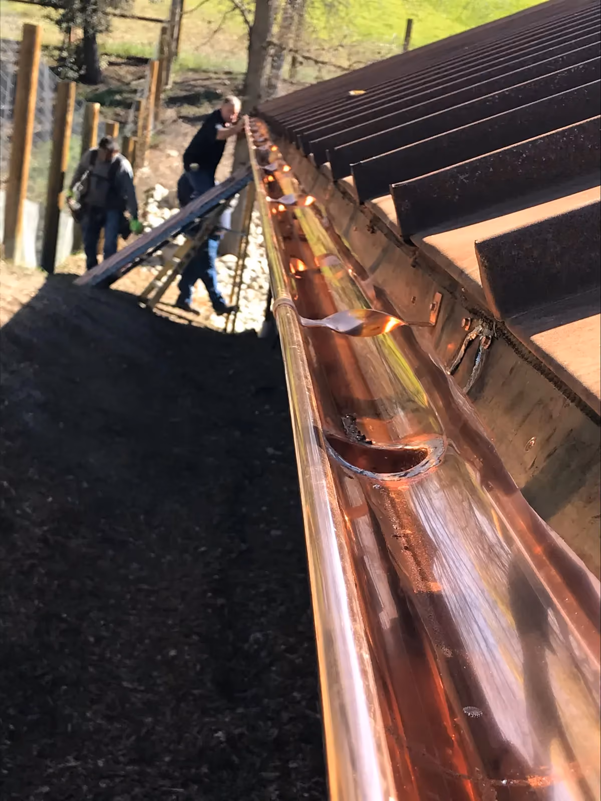 A close view of a copper gutter along a stripped roof deck, a crew member and a ladder behind it.