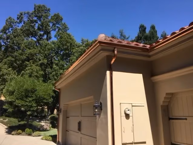 A finished copper gutter and downspout along the eave of a stucco house with a clay tile roof, oaks and blue sky behind it.