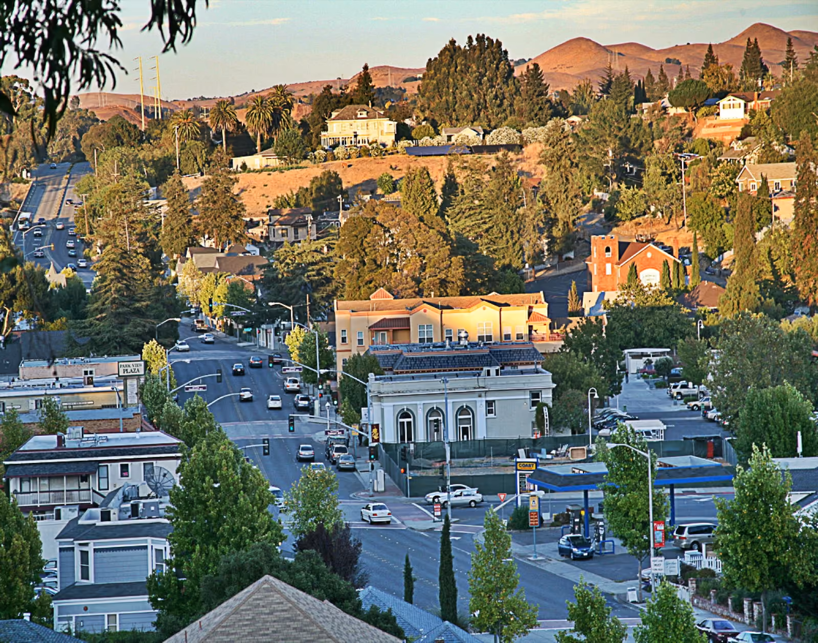A main street of low storefronts from above, wooded hills and houses climbing behind it.