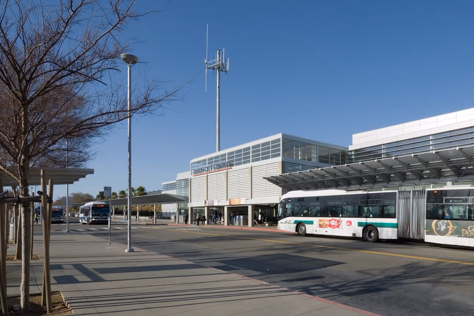 A long station canopy with a bus waiting beneath it and bare trees along the kerb.